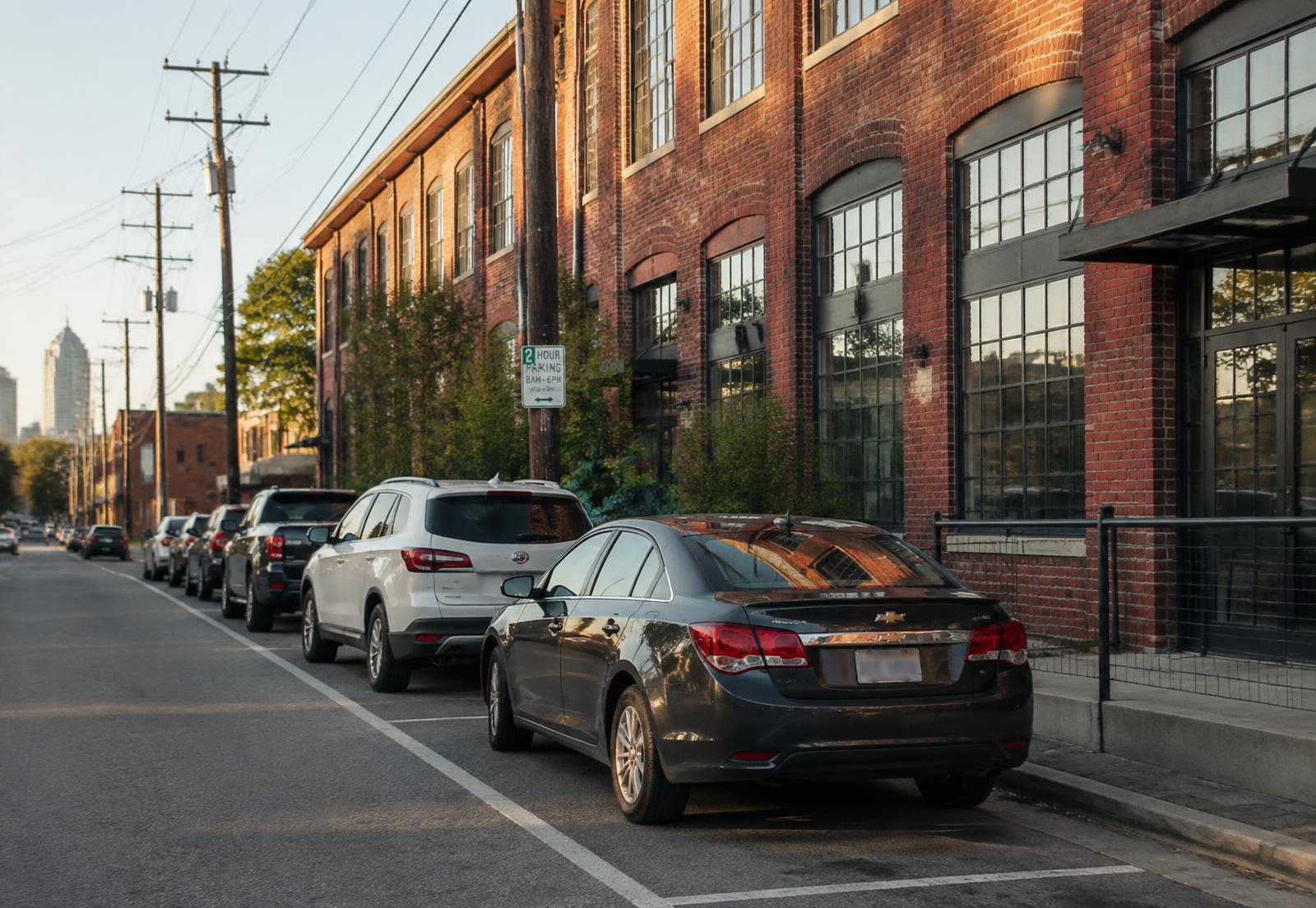 Cars parked along a brick street beside a converted red brick mill building in NoDa, Charlotte, at golden hour